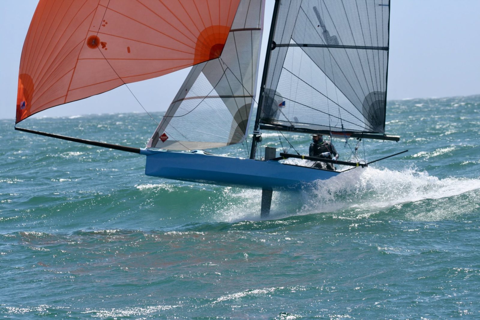 An I-14 skiff fully powered up on foils under an orange asymmetric spinnaker, throwing spray